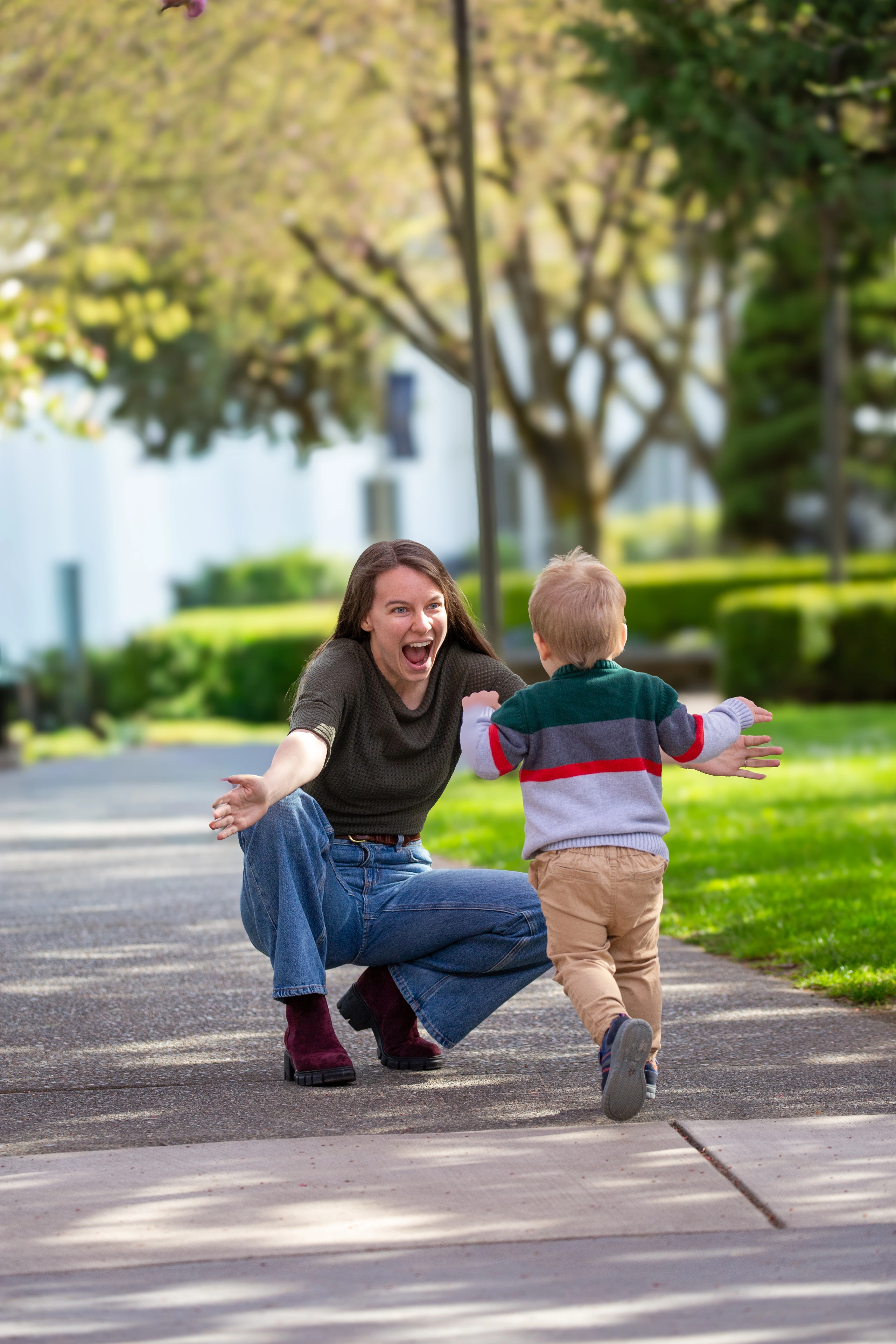 A mother kneels with open arms as her young child runs toward her beneath spring trees.