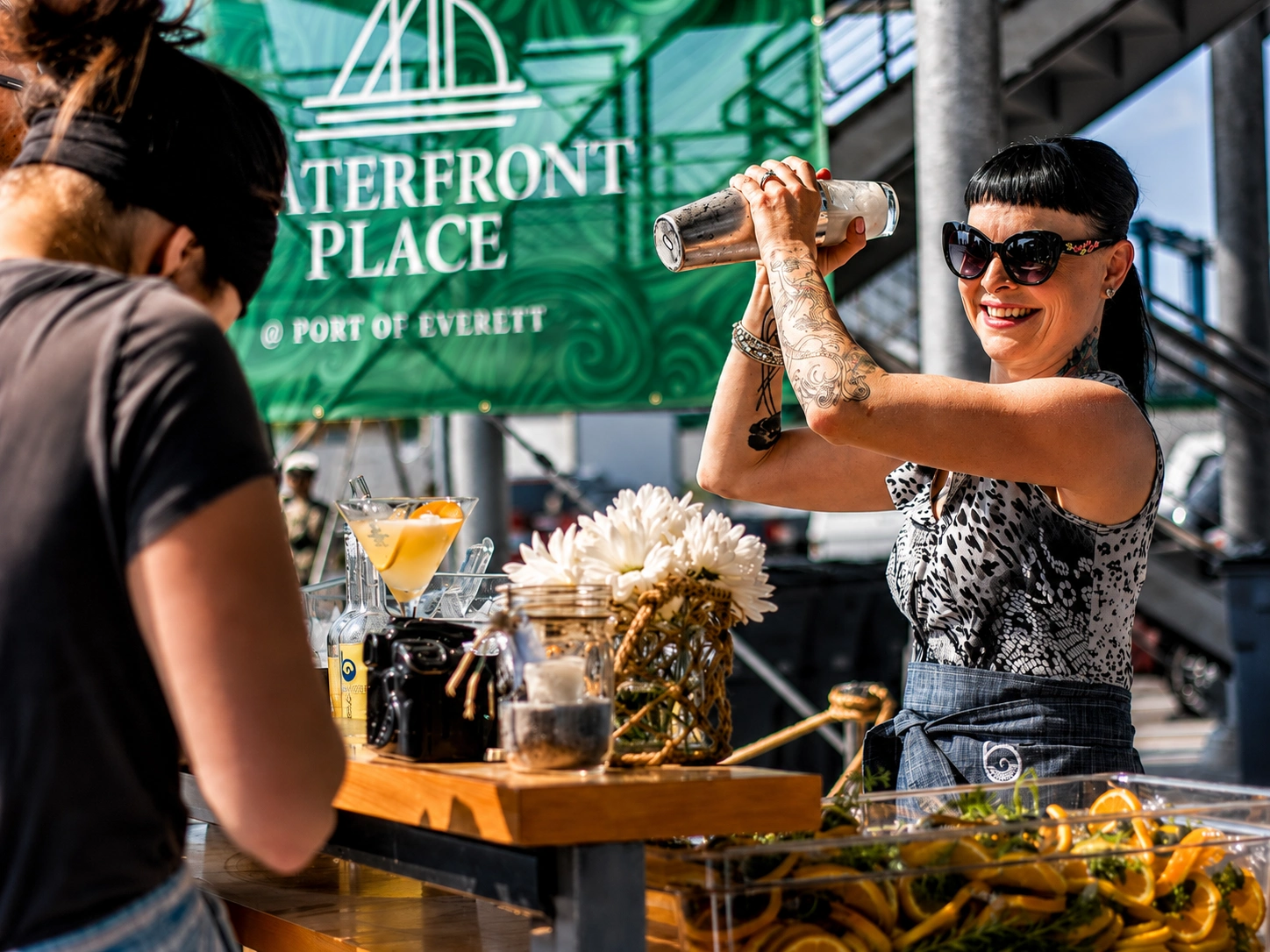 A smiling bartender shakes a cocktail at an outdoor event.