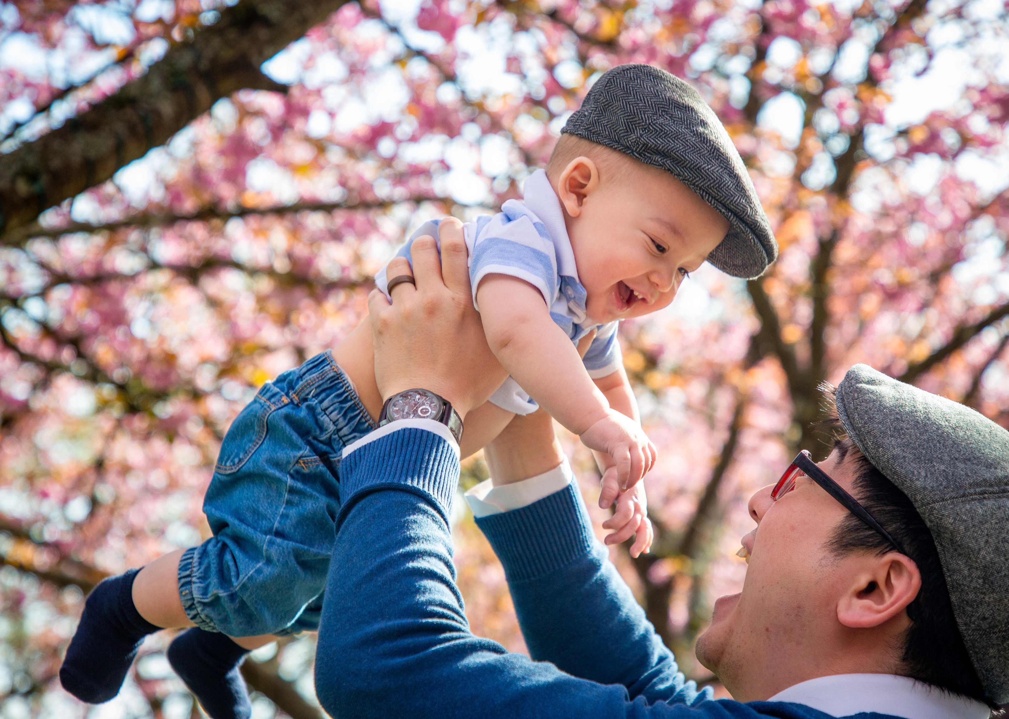 A father lifts his laughing baby into the air beneath blooming pink branches.